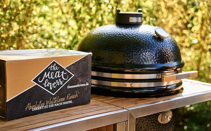 ein-karton-mit-fleisch-von-the-meatlovers-und-daneben-ein-monolith-keramikgrill